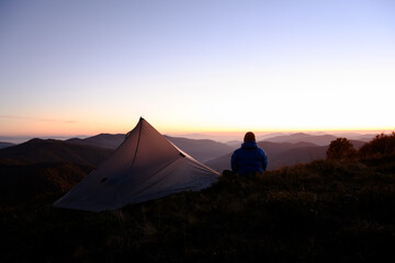 Traveler near tent in highlands framed by stunning autumn sunset and mountain peaks. Scenic outdoor silhouette perfect for golden hour landscape moments