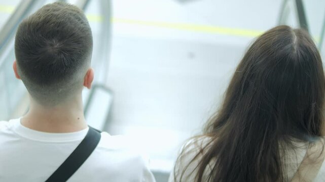 Man and a woman are standing next to each other on an escalator. The man is wearing a white shirt and a black backpack. The woman has long brown hair
