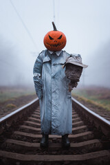 Spooky figure in carved pumpkin mask with scarecrow severed head in hand stands on misty railroad in autumn countryside. Halloween concept