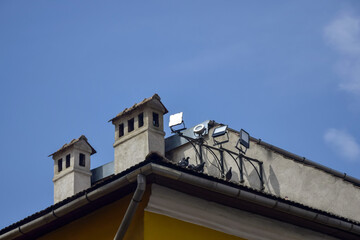Floodlights installed on roof of old building with ventilation columns and pipes for rainwater drainage. City pigeons have taken fancy to  roof. Against backdrop of clear blue sky. Copy space for text