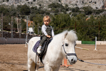 Young child rides a white pony in an outdoor riding arena surrounded by scenic nature