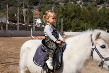 Child riding a white pony in a riding arena on a sunny day