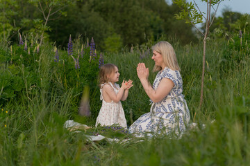 A happy mom and little daughter are sitting on a blanket in the park, clapping their hands. A happy...