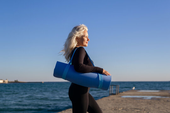 Yoga Mat Woman Beach Exercise - A woman in black athletic wear carries a blue yoga mat on her way to exercise on the beach.