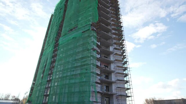 Unfinished skyscraper stands wrapped in fluttering green tarps under sky. Steel beams and concrete slabs remain exposed on upper floors
