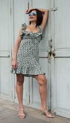 Stylish Asian woman leans confidently against a weathered door, wearing a trendy floral-patterned dress paired with beige high-heeled sandals