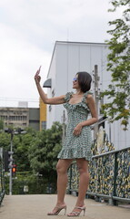 Modern Asian woman dressed in a green floral dress with ruffled straps and strappy heels. She takes a selfie with a pink phone and wears blue-lensed sunglasses