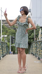 Confident Asian woman dressed in a green floral dress with ruffled straps and strappy heels. She takes a selfie with a pink phone and wears blue-lensed sunglasses