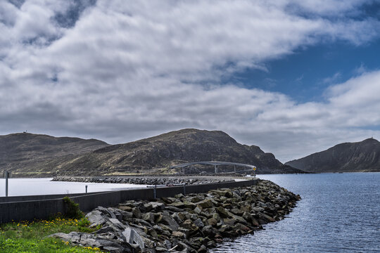Scenic Coastal Road Bridge Next to Calm Waters and Rocky Terrain Under Cloudy Sky, Runde, Norway