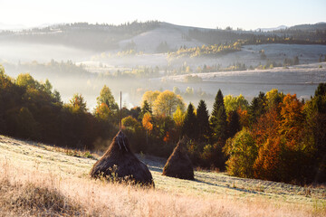 Foggy pasture with haystacks and orange forest glows in sunrise light in autumn mountains. Serene highland landscape with dried hay and foggy hills