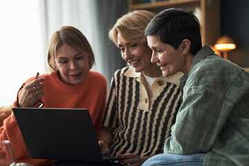 Three middle aged Caucasian women sitting closely together using laptop, smiling and interacting, demonstrating friendship and connection in casual indoor setting