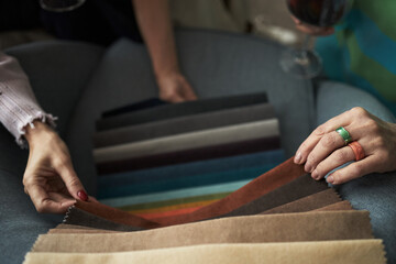 Two young adult Caucasian women selecting fabric samples together, hands holding swatches in various colors, one woman holding wine glass, engaging in creative decision making