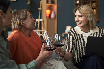 Three middle aged Caucasian women sitting together smiling and holding wine glasses, enjoying friendly conversation in cozy living room setting, celebrating adult friendship