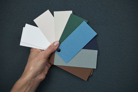 Caucasian young adult woman hand holding color swatches against dark textured wall, displaying various shades of paper samples in fan arrangement for design or decoration concept