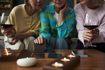 Three Caucasian middle aged women sitting together holding glasses of red wine, engaging in tarot card reading at table with candles, focusing on cards and conversation