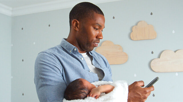 Father Holding Newborn Baby Son Whilst Using Mobile Phone In Nursery At Home