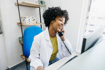 Smiling medical receptionist talking on phone and using computer, managing appointments and...