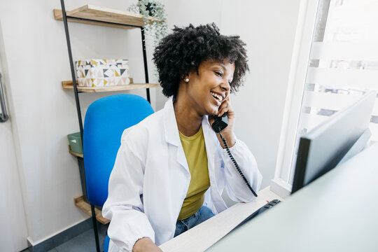 Smiling medical receptionist talking on phone and using computer, managing appointments and providing customer service in healthcare clinic