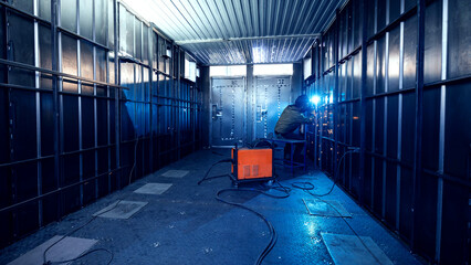 Caucasian male welding in industrial factory room with metal walls and equipment, blue light