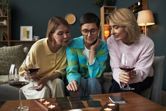 Three middle aged Caucasian women sitting together on sofa holding glasses of red wine, engaging in tarot card reading with lit candles on table, smiling and interacting closely