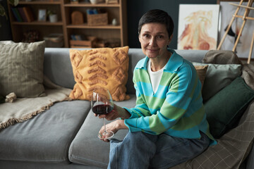 Portrait of middle aged Caucasian woman sitting on sofa holding glass of red wine, short dark hair, relaxed facial expression, looking into camera, casual home setting
