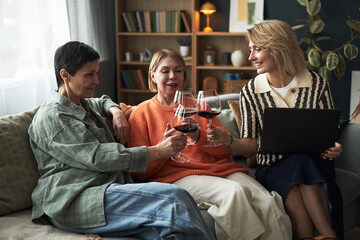 Three Caucasian middle aged women sitting on sofa clinking wine glasses, smiling and engaging in conversation, one holding laptop, casual indoor setting with bookshelves in background