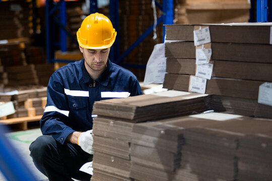 Man paper warehouse worker using clipboard checking inspecting quality cardboard in storage. Large industry in paper product line manufacturing. Employee level wear uniform and helmet yellow.