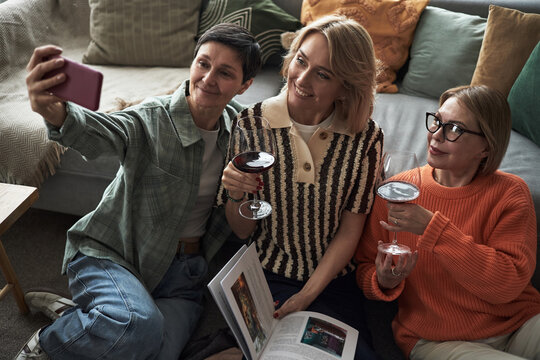 Three Caucasian middle aged women sitting on floor taking selfie with smartphone while holding wine glasses and open book, smiling and looking at phone together - Powered by Adobe