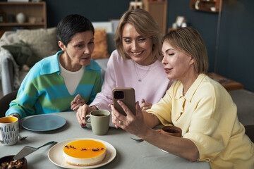 Three middle aged Caucasian women sitting at table smiling and looking at smartphone together, enjoying dessert and drinks, showing close friendship and relaxed interaction