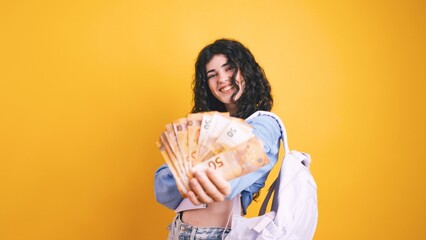 Happy student showing money and holding backpack on yellow background