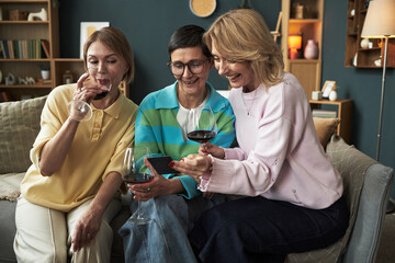 Three middle aged Caucasian women sitting on sofa smiling and drinking wine, one woman holding smartphone showing something to friends, all appearing relaxed and enjoying time together