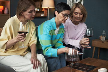 Three middle aged Caucasian women sitting together holding wine glasses, playing card game on table, focusing on cards, engaging in social activity in cozy indoor setting