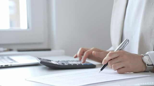 Businesswoman is calculating annual tax using calculator and taking notes in the office, managing domestic budget and checking financial documents. Audit and taxes in business