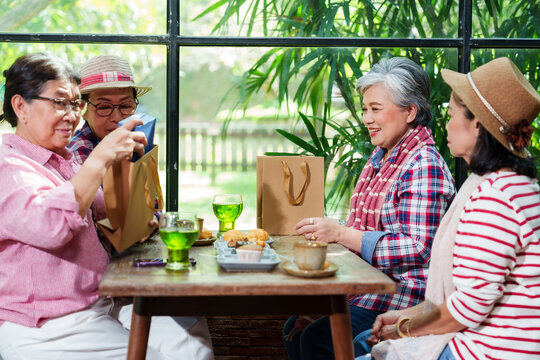 Group of senior Asian women exchanging gifts sitting cafe enjoying relaxing cheerful casual colorful clothing smiling conversation friendly bonding indoor natural greenery background leisure