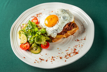 Crispy waffle topped with pulled pork, melted cheese, and sunny-side-up egg, served with fresh salad of cherry tomatoes and cucumber slices on a white plate.