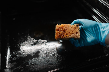 Male hand wearing blue rubber glove holding dirty sponge, scrubbing soiled gas oven covered in...