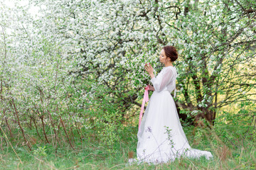 A young woman in a white dress stands among flowering trees, enjoying the spring nature.