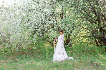 A young girl in a white dress stands among flowering trees, surrounded by greenery. Her gaze is...