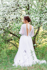 A girl in a white wedding dress with an open back stands against the background of blossoming...