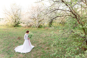 A beautiful girl in a white dress stands among blossoming trees, surrounded by spring greenery....
