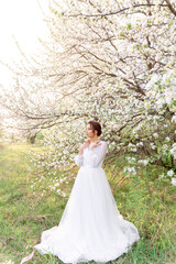 A girl in a white dress stands under blossoming trees, surrounded by greenery. Bright morning,...