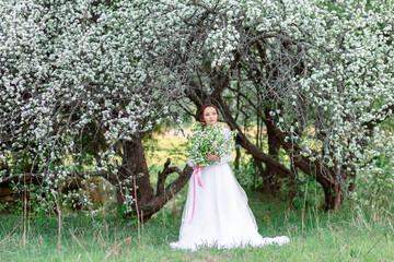 A young girl in a white dress stands under blossoming trees, holding a bouquet. Spring landscape,...