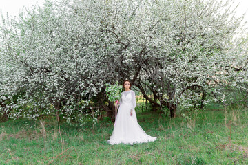 The bride in a white dress stands under blossoming apple trees, surrounded by spring greenery,...