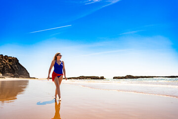 Beautiful middle aged woman walking on sandy beach in summertime. Front view. Monte Clerigo beach on Algarve coast in Portugal