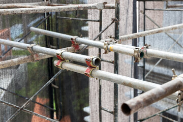 Close-up view of metal scaffolding pipes and couplers at a construction site, showcasing the structural connections and weathered textures. The image emphasizes industrial materials, safety equipment,
