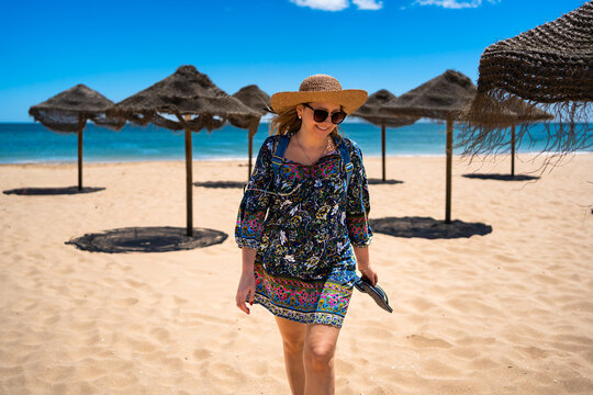 Beautiful happy middle aged woman tourist in straw hat walking on sandy beach full of beach umbrellas in summertime. Front view. Meia Praia beach in Lagos, Portugal