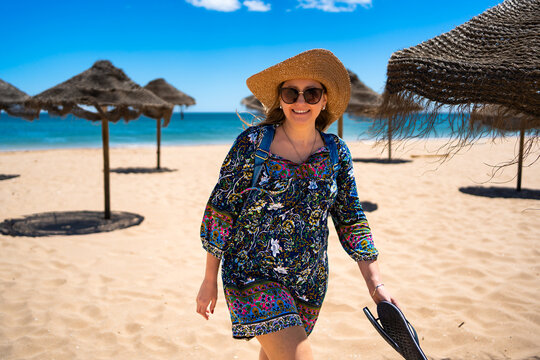 Beautiful happy middle aged woman tourist in straw hat walking on sandy beach full of beach umbrellas in summertime. Front view. Meia Praia beach in Lagos, Portugal - Powered by Adobe