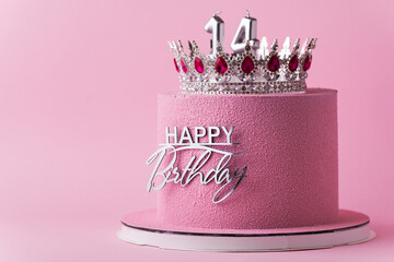 Beautiful luxury birthday anniversary cake for a girl with pink chocolate velvet coating decorated with silver crown and happy birthday topper. Pink background. Happy 14 years old celebration concept