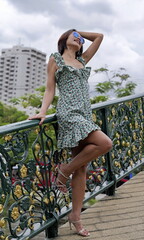 Stylish Asian woman leans on a decorative bridge with green and gold railings, wearing a floral dress with ruffled straps and strappy heels