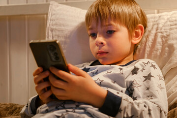 Cute kid boy in pajamas lying in bed watching smartphone, smiling. Children's screen time. Child plays games using phone at the evening or night. Social media addiction. Childhood, modern technology